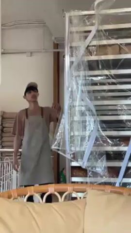 A young man in a cafe or bakery wearing a hat, T-shirt, and apron retrieves a tray of baked goods from a shelf above a doorway. The shelves appear to be full of loaves of bread in wrappers, some kind of sourdough bread. He carefully removes the tray and carries it off to the left, away from the camera. To his right is a rack of baking trays covered in plastic. The space appears to be light and airy with a relaxed decor and a sign with the Wi-Fi information.