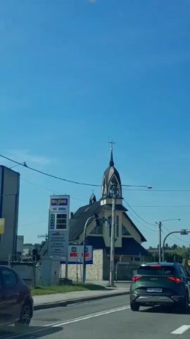 The video shows a street scene on a clear day with a blue sky. A church with a tall, dark steeple topped with a cross is visible in the background. To the left of the church is a gas station sign with prices displayed. A green Kia Sportage is driving on the road in the foreground, followed by an orange car. Several streetlights and traffic signals are present. A few people are standing on the sidewalk in front of the church. The camera appears to be moving, possibly from inside another vehicle.