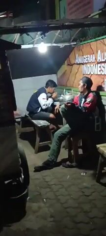 Two young men are seated at separate low wooden benches under a covered outdoor area at night. The man on the left, wearing a dark blue and white jacket, is leaning forward and appears to be talking to the man on the right. The man on the right, wearing a red and black jacket and green pants, is seated with his legs crossed and is looking towards the man on the left. Between them, on a table or cart, is a metallic container, possibly a teapot or bowl, and other items. A bright light bulb hangs overhead, illuminating the immediate area. Behind the men, a sign with text in Indonesian is visible, including the words "ANUGERAH ALAM INDONESIA". To the left of the men, part of a food cart or stall is visible, displaying the text "RAKSO LAYAT MENU BAKSO" along with images of food. The ground is paved with interlocking bricks. In the background, a street is dimly lit by distant lights, suggesting a roadside setting. The overall scene appears to be a casual gathering at a food stall during the evening.