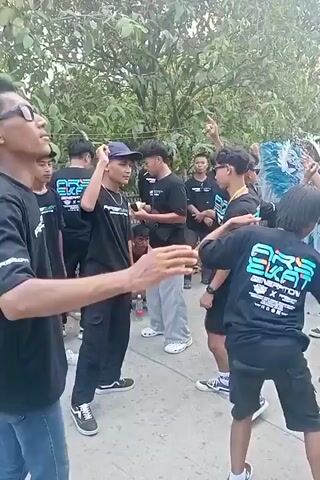 The video shows a group of young people gathered outdoors. They appear to be in a casual setting, possibly a street or a park, with trees and foliage in the background. Most of the individuals are wearing black t-shirts with a teal and white logo that says "ARS SKAT GENERATION". Some are wearing baseball caps.

In the foreground, a person in a black t-shirt and blue jeans faces the camera and gestures with their hands. To their left, another person in a black t-shirt and black pants, wearing a blue baseball cap, is looking towards the center of the group and appears to be talking or interacting with others.

In the center of the group, several individuals in black t-shirts are interacting. One person holds a small white object. Another person, wearing light grey pants and white Crocs, is looking towards the left. Two individuals in the foreground on the right are facing away from the camera, one with a black t-shirt and shorts, and the other with a black t-shirt and pants. The person in black shorts is wearing white and black sneakers. The person in black pants is wearing dark sandals.

The overall atmosphere appears to be one of casual gathering and socializing, with some individuals engaged in energetic movements or poses. There's a general sense of youth culture and camaraderie among the group.