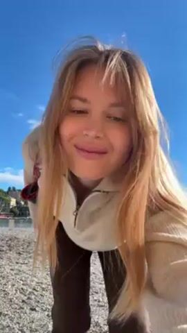 A woman stands on a pebbled beach under a bright blue sky with scattered clouds. She is facing the camera, then straightens up, placing her hands on her hips. She is wearing a light beige sweater, dark brown pants, and shoes of similar tone to the sweater. She has shoulder-length blond hair with bangs. A black purse with a red strap hangs from her shoulder. In the background, across the water, is a town with buildings and trees, and distant mountains.