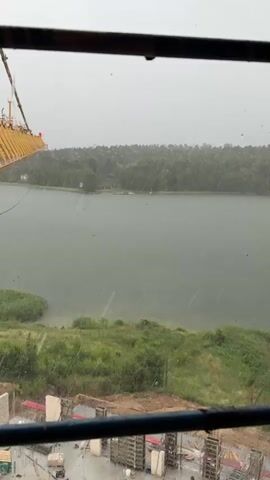 The video shows a view from inside a building, looking out at a body of water and surrounding greenery. Rain is falling, visible as streaks across the glass. In the foreground, there is a construction site with concrete structures and scaffolding. A yellow crane is visible on the left side of the frame, extending over the water. The water appears calm, with trees lining the far bank. The sky is overcast.