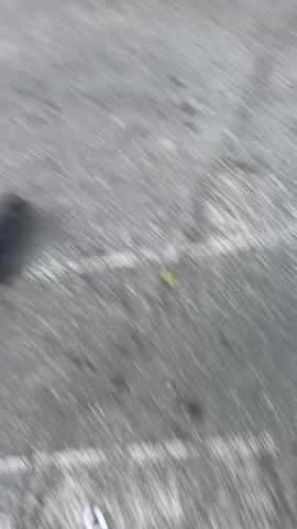 A person's legs and feet are visible in the video, wearing black shoes and dark pants. The individual appears to be walking or running on a gray asphalt surface, possibly a road or parking lot. The camera angle is low, focusing on the lower extremities and the ground. There is some debris on the asphalt, including leaves. The motion blur suggests movement.