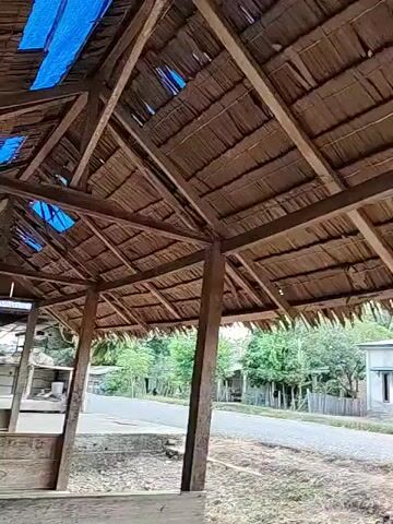 The camera pans slowly across the scene from under a simple structure with a thatched roof made of thin, flat branches.  The supports of the structure appear to be made of roughly hewn wood.  Outside the structure, the scene consists of a few simple houses, a paved road, and various trees and green foliage. The ground near the structure is rough, with loose rocks and dirt.  The lighting suggests it is daytime, possibly late afternoon.  The sky is overcast.