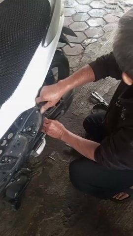The video depicts a person, appearing to be an older male with short gray hair, squatting next to a white and black scooter. The person is wearing a dark brown long-sleeved shirt, black pants, and dark sandals. They are focused on the area beneath the seat and side panel of the scooter, seemingly working with their hands and possibly a small tool to detach or adjust something. Various tools, including a plier-like tool and a socket wrench, are scattered on the gray and red brick-patterned ground around them. The scooter is parked on the brick surface, with its rear tire visible in the background. The overall environment suggests an outdoor or semi-outdoor workspace, potentially a garage or driveway.