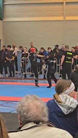 Here's a detailed description of the video:

The video shows two young people sparring in what looks like a martial arts competition. They are in full gear, including headgear, gloves, and shin guards. They are on a blue and red mat in the center of a gymnasium. 

Around the mat is a crowd of spectators standing behind a black metal barricade. There are people of various ages and genders. Several appear to be coaches or staff, with one person wearing a black shirt that says "STAFF" and "TEAM ORIGLIA" on the back.

A referee in a dark green top stands near the sparring athletes, observing their movements and ensuring the rules are followed. The fighters exchange punches and kicks, with the referee occasionally intervening. The crowd watches intently, some cheering. The environment is indoors with bright lighting, typical of a gymnasium. The camera is held by someone in the audience, who does not move much during the recording.