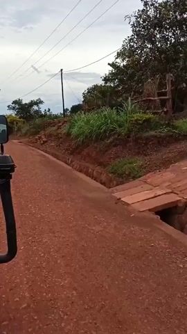 The video depicts a vehicle moving along a red dirt road. The road is lined with shallow, concrete ditches on either side, and the adjacent land is covered with vegetation, trees, and some exposed reddish soil and rocks. Power lines are visible overhead against an overcast sky. The overall impression is of a rural, possibly tropical location.