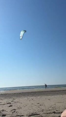 A lone figure stands on a sandy beach, silhouetted against the vast expanse of the ocean and a clear blue sky. The person is kiteboarding, holding a kite in the air. The kite is large and white with green and red accents, and it appears to be catching the wind. The waves are gentle, lapping at the shore. The sun is high in the sky, suggesting it is midday. The beach is mostly empty, with a few footprints in the sand. The overall scene is peaceful and serene, with a sense of freedom and adventure. The location appears to be Petten, Netherlands, given the context provided. There is no visible text in the media file.