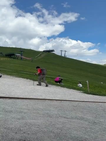 This is a vertical video file, shot outdoors on a sunny day with scattered clouds. The scene depicts a mountain landscape with rolling green hills dotted with yellow wildflowers. In the background, jagged, rocky mountain peaks dominate the horizon, bathed in sunlight. A ski lift system is visible on the hillside, with its towers and cables extending upwards.

In the foreground, several people are present. A man in a red shirt and backpack is walking with a hiking pole. Further up the hill, two individuals are seen. One, wearing a pink backpack, is crouching, and another person is sitting beside them. As the camera pans to the right, more people are visible scattered across the grassy slopes, walking along a winding path. A person in a teal shirt stands facing the mountains, observing the vista. The terrain in the immediate foreground is a gravel path.

The overall atmosphere suggests a leisurely day spent outdoors, likely a hiking or scenic viewpoint location, possibly at a ski resort during the off-season, given the presence of the ski lift. The bright sunlight and clear blue sky indicate pleasant weather, ideal for outdoor activities and enjoying the natural beauty of the mountainous region of Santa Cristina in Gröden, Italy. There is no visible text in the video.