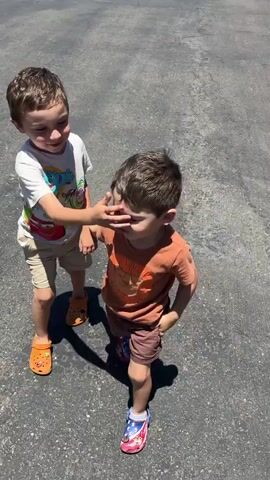 Two young boys are walking hand-in-hand on an asphalt surface. The boy on the left is wearing a white t-shirt with colorful graphics and tan shorts. He has orange crocs on his feet. The boy on the right is wearing an orange t-shirt with a lion graphic and brown shorts. He has red, white, and blue crocs on his feet. The background shows a parking lot with parked cars under a covered structure. The sun is shining, casting shadows.