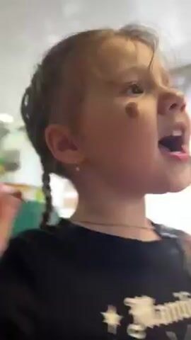 A young child with light hair is the main focus of the video. The child wears a black shirt with a white design. They are holding a yellow and a green object in their hands and moving them up and down while looking upwards and talking. The child has a mole on their left cheek. The background shows a room with a window covered by a curtain and a television on the wall. There's also a picture of a man with a beard and glasses on the wall. Another person with light hair and wearing a black shirt is briefly visible in the background.