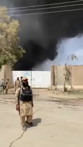 A group of armed soldiers stand outside a compound in a Middle Eastern or Central Asian country. Behind them, massive plumes of black smoke rise into the sky, suggesting a fire. The soldiers are dressed in beige-colored camouflage uniforms. The soldier in the foreground is wearing a tactical vest and has a beard. The compound's entrance is a white gate with a sign bearing the blue and white logo of a company called "SAIPEM." The environment appears dry and dusty with sparse vegetation.