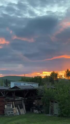 The video captures a picturesque sunset scene. In the foreground, a wooden shed is visible, surrounded by lush greenery and trees. Behind the shed, a white building with a blue stripe is partially visible. The sky is dominated by a dramatic cloudscape, with grey clouds tinged with orange and pink hues from the setting sun. The sun itself is not directly visible but its light casts a warm glow on the horizon. The overall scene is tranquil and serene, showcasing the natural beauty of the sunset against the backdrop of a rural landscape. The camera is static throughout the video, providing a fixed view of the scene.