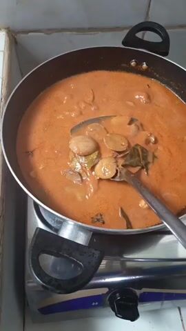 A close-up, high-angle shot shows a dark-colored pot of stew or curry being cooked on a gas stove. The stew is a reddish-orange color and appears to contain pieces of meat or vegetables, possibly jengkol. A ladle is being used to lift some of the stew and its contents from the pot. The pot has black handles and is sitting on a silver and black gas stove with a blue trim. The background is out of focus, but appears to be a tiled wall.