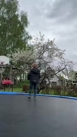 A young boy, possibly around four years old, is seen on a trampoline in an outdoor setting. He is wearing a dark jacket, green pants, and black sneakers. The boy walks forward, faces the camera, smiles, and then briefly jumps on the trampoline. Behind him, there's a landscape featuring trees with white blossoms, green shrubbery, and a grey house in the background. The sky is overcast. A basketball hoop and other playground equipment are visible to the left.