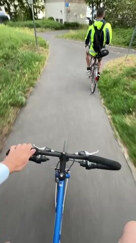 The video shows a first-person perspective of someone riding a blue bicycle, following another person on a bicycle from behind. The lead cyclist, wearing a neon yellow jacket and a black backpack, is riding a red bicycle and makes a left turn from an asphalt path onto a cobblestone path. The scene is outdoors on a paved path lined with green grass and trees. In the background, buildings are visible, including one with a blue and white 'P' parking sign. The sky is partly cloudy. The setting appears to be an urban or suburban area in Hattersheim am Main, Germany, during the daytime.