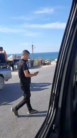 The video shows a man walking on a road, looking at his phone. He is wearing a black t-shirt and black pants. In the background, there are some stalls or shops, and the sea can be seen in the distance. A white truck is driving on the road. The sky is blue with some clouds.