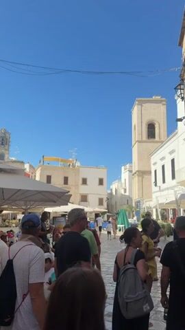A group of people are walking down a narrow street lined with buildings and outdoor seating areas. The sky is clear and blue, and the sun is shining brightly. One building has a tower with an arched opening. Another building has a balcony with potted plants. People are dressed in casual summer clothing. Some people are carrying backpacks. There are umbrellas providing shade over tables.