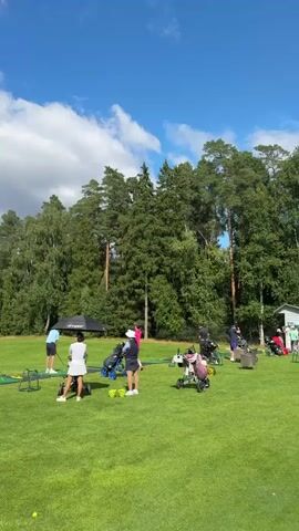 A group of people are on a golf course practicing. Some are hitting golf balls from mats with golf clubs, while others are standing and watching. There are golf bags and equipment set up on the grass. The environment is a grassy field with trees in the background and a blue sky with clouds above. The weather appears to be sunny.