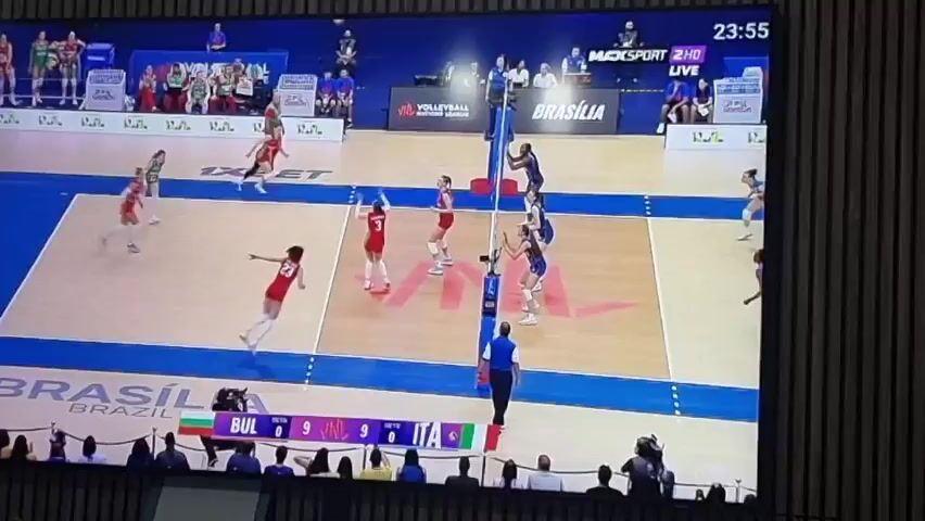 The media file displays an indoor women's volleyball match in progress. Two teams are featured: one in red and green uniforms, identified as Bulgaria (BUL) by the flag on the scoreboard, and the other in blue uniforms, identified as Italy (ITA) by its flag.

The scoreboard clearly shows the current score as Bulgaria 0 sets, 9 points, and Italy 0 sets, 9 points, indicating a tie during a set. A digital clock in the top right corner of the screen displays 23:55.

The video shows a sequence of play. In the initial frame, a player in a blue uniform is seen on the court floor, having just made a defensive maneuver. Other players are positioned around the net. The scene then cuts to three players from the red team (Bulgaria) high-fiving each other near the net, celebrating a point or a successful play.

The volleyball court is brightly lit and features distinct blue and light brown sections. The words "VOLLEYBALL" and "BRASÍLIA" are visible painted on the court, suggesting the tournament or event's location or name. Spectators are seated in the stands surrounding the court in the background. A large screen in the arena displays "MACKSPORT 2HD LIVE" in the upper right corner, indicating a live broadcast.