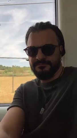 The video is a selfie-style shot of a man with dark hair, a beard, and sunglasses, wearing a dark t-shirt. He is sitting inside a vehicle, possibly a train, as indicated by the window and the passing scenery of fields and trees. He has earbuds in and a pendant necklace. He turns his head slightly to the right.