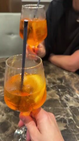 The video shows a marble table with three glasses of Aperol Spritz. The glasses are filled with ice, an orange slice, and a black straw. Two hands are holding the glasses, bringing them together for a toast. People are visible in the background.