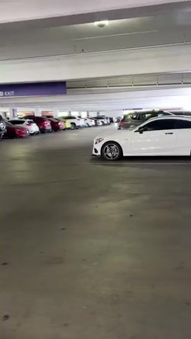 The video shows a panning view of a parking garage. The camera moves from left to right, capturing several cars parked in the garage. The cars are of various makes and models, and some are white, black, or red. The environment appears to be well-lit with overhead lighting. There are signs visible that say "EXIT" with purple and green backgrounds. The floor of the parking garage is concrete. The video does not show any people or any interactions. The perspective suggests the camera is being held by someone walking or moving through the garage.
