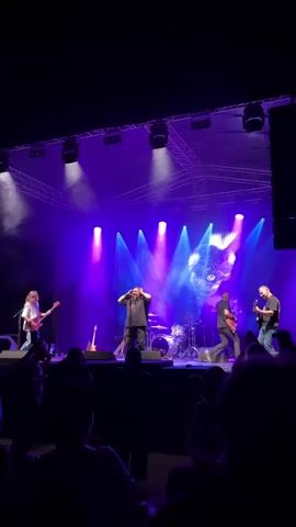 A live band performs on a brightly lit stage, illuminated by purple and white spotlights. The band consists of a lead singer in the center, wearing a dark shirt and pants, with their hands behind their head. To the left, a guitarist with long hair plays a white guitar. To the right, another guitarist plays a guitar. A drummer is visible behind the lead singer, seated at a drum kit. The stage backdrop features a geometric light projection, possibly of a face or abstract design, and there are musical instruments, including guitars, on stands. The audience, silhouetted in the foreground, watches the performance. The atmosphere appears to be energetic, suggested by the stage lighting and the performance itself.