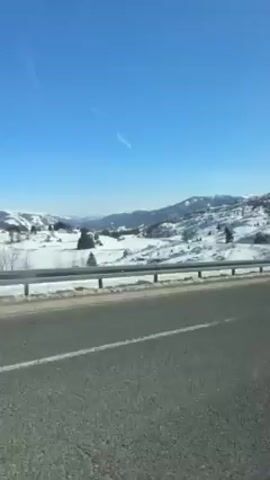The video shows a snowy landscape seen from a car window as it drives along a highway on a sunny day. The road is paved and has white lane markers. A metal guardrail separates the road from the snow-covered field beyond. In the distance, snow-covered hills and mountains can be seen under a clear, bright blue sky.  The video is shot from inside the vehicle and the window reflects some light.