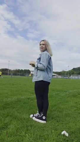 A young woman with blonde hair, styled with pink streaks, stands on a grassy field. She is wearing a denim jacket, black leggings, and high-top black and white sneakers. Her body is angled to the right, and she is looking slightly to her left, with her hands near her chest. In the background, several people in athletic wear are visible on what appears to be a soccer field, with goals and stadium lights in the distance. The sky above is partly cloudy with patches of blue.