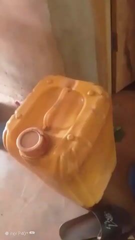 The media file shows a yellow plastic jerrycan with a red cap placed on a floor. The jerrycan is large and appears to be filled with a liquid. It has a handle on top and a spout. To the left of the jerrycan, there is a dark object, possibly a pipe or a container. In front of the jerrycan, a pair of black flip-flops with white accents are visible. The background consists of a light-colored wall, suggesting an indoor setting. The lighting is dim, indicating it might be daytime with indirect light, or evening. The image is slightly blurry, particularly at the edges and in the background, which is characteristic of a photo taken with a mobile phone. The camera watermark at the bottom left corner reads "©itel P40+". The scene appears to be a domestic or storage area, possibly in Achara Ndiagu Omegio, Nigeria, given the location context. No people are visible in the media file. The overall impression is a still life of everyday objects in a simple setting.