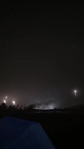 The video shows a nighttime scene with fireworks. The first few seconds capture bursts of white and pink fireworks against a dark sky. Then, the scene transitions to a deep red hue, suggesting a large, bright red light source is illuminating the sky and clouds, possibly another firework or a strong spotlight. The video ends with a shot of the red illuminated sky. In the foreground, the silhouette of a dark, possibly tent-like structure is visible. In the distance, faint lights of a settlement or town are discernible below the fireworks and the red glow.
