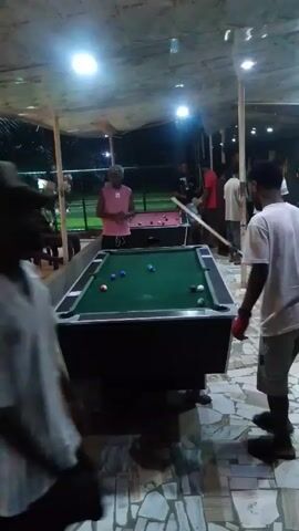 Here is a description of the video:
A group of people are gathered under a covered patio, illuminated by overhead lights. They are playing pool on two tables. The camera focuses on one man, dressed in a white shirt and gray shorts, who is lining up a shot on a pool table.  A man in a pink shirt watches him. Other people are standing around the pool tables. Beyond the patio area, there is a glimpse of green foliage. The camera is handheld.