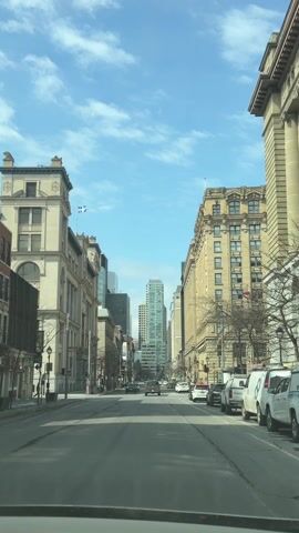 The view from the interior of a vehicle looks down a straight city street lined with tall, architecturally detailed buildings, predominantly tan in color. A few modern, glass skyscrapers are visible in the distance. The sky is bright blue with scattered white clouds. Several cars drive down the street while others are parked along the right side. A single pedestrian walks on the left sidewalk. Traffic lights are visible at intersections, and the Quebec flag flies from one of the buildings on the left.