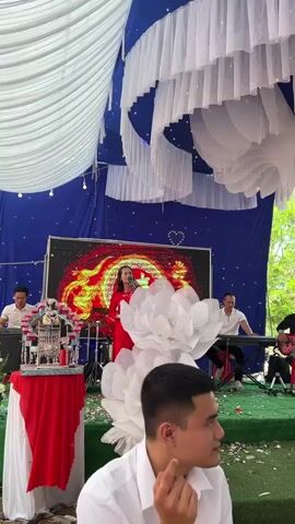 The video captures an outdoor celebratory event, likely a wedding or a festive gathering, taking place under a large, decorated tent in Tay Son Commune, Vietnam. The tent's ceiling is elaborately draped with white and blue fabric, interspersed with string lights, creating a bright and festive atmosphere.

At the front of the tent, a stage is set up for live music. A female singer, dressed in a striking red gown, is performing into a microphone. Behind her, a large LED screen displays vibrant, abstract patterns resembling fiery light. A band accompanies the singer, consisting of a keyboardist on the right and a guitarist next to him, with a drummer partially visible further back. The stage is decorated with prominent, multi-layered white floral structures, resembling large lotus blooms.

Guests are visible throughout the tent, either seated at tables covered with red tablecloths or standing and mingling. In the foreground, a man in a white collared shirt is seen looking towards the right. As the camera pans slightly, two young women also appear in the foreground, attentively observing the event. Towards the end of the clip, a man in a dark suit and a woman in a bright yellow dress are seen walking through the seated guests, with what appears to be a bride in a white gown partially visible behind them. The tent's side sections are adorned with red and white fabric drapes, enhancing the celebratory decor. The overall scene suggests a lively and joyous occasion.