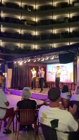 A group of three young men in matching white shirts and black pants are performing on a stage with a purple-lit floor and a large screen behind them. They are singing and gesturing, with one of them holding a microphone. Several audience members are seated at tables in the foreground, watching the performance. The stage is set up outdoors, possibly at a resort, with a multi-story building visible in the background, illuminated by rows of lights. The overall atmosphere appears to be entertainment and leisure.