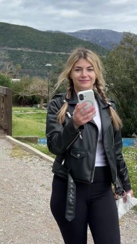 A woman in a leather jacket, white shirt, and pigtails stands in a park, looking off-camera and holding a phone. The camera pans to a white and brown Jack Russell terrier with a short tail, wearing a red collar. The dog is standing on a gravel path near a small wooden bridge. Someone off-camera says "Иди сюда", which means "Come here" in Russian, and the dog walks toward the camera.