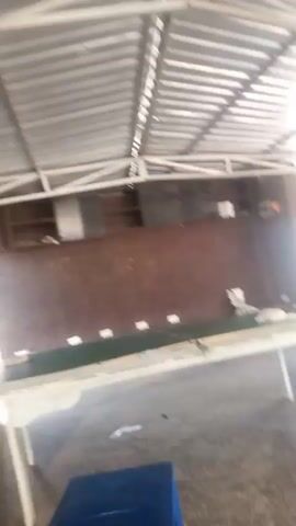 The video shows an indoor scene with a corrugated metal roof supported by white beams. A long, white table with a dark green surface is positioned in the center of the frame. Several white objects are placed along the table. A dark reddish-brown wall is visible behind the table, with shelves and some indistinct objects on them. The floor appears to be concrete. In the foreground, a blue object is partially visible, and to the left, a stack of gold-colored cylindrical objects, possibly drums, are present. The lighting is bright, suggesting daylight. The camera is moving and the footage is somewhat blurry.