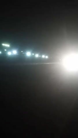 A low-angle night shot shows a truck approaching and then passing on a dark road. The truck's bright headlights illuminate the road as it gets closer. As it passes, the red and amber lights on the back and top of the truck become visible before it drives out of frame. The sky is dark, and several bright lights are visible in the distance on the left side of the frame, suggesting a nearby town or other development.