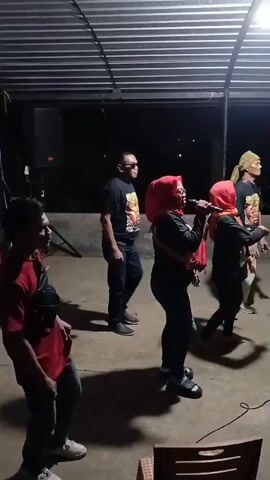 The video shows a group of five people in what appears to be an outdoor covered area at night. The ground is paved. In the background, there are dark trees and the sky. A metal roof structure is visible above. On the left, a person wearing a red t-shirt and jeans stands facing left, with a black bag slung across their chest. Next to them, to the right, a man in a black t-shirt with a graphic print and jeans is dancing. He is wearing sunglasses and has his left arm extended, pointing. To the right of him, a woman in a red hijab and black clothing is singing into a microphone. She is also wearing a red scarf around her neck. To her right, another person in a black hijab and black clothing is dancing and has their right arm raised. Further to the right, a person wearing a yellow head covering and a black t-shirt with a graphic print is standing and watching. There is a speaker on a stand to the far left of the frame. A dark electrical cord runs along the ground in the foreground. A wicker chair is partially visible at the bottom right. The lighting is dim, with some artificial light sources contributing to the scene. The people appear to be engaged in a performance or social gathering.