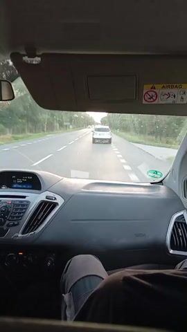 The video shows the interior of a vehicle from the perspective of a passenger. The dashboard is visible, with a navigation screen displaying information. The road ahead can be seen through the windshield, with another white car driving in front. Trees and greenery line the sides of the road. The sun appears to be setting, casting a warm light. A person's legs and lower body are visible in the foreground, suggesting they are sitting in the passenger seat. The overall impression is a leisurely drive in the countryside.