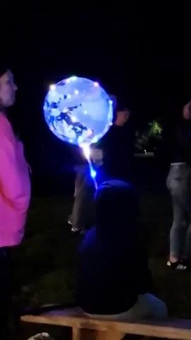 The video shows a group of people at night in an outdoor setting. One person, whose face is not visible, is holding a large, translucent blue balloon that is illuminated by small, multi-colored lights. The balloon appears to have a dark pattern or image on its surface. The person holding the balloon has a bright blue light emanating from what appears to be the balloon's stem, which is connected to a stick or handle. Several other people are visible in the background, most of them partially obscured or out of focus. One person on the left is wearing a bright pink top. The ground appears to be grass or dirt. The overall atmosphere is dark, suggesting it is nighttime. The people seem to be gathered around, possibly observing the balloon.