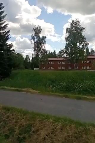 This video shows a view from a moving vehicle, likely a car or bus, on a paved road. The camera pans from left to right, revealing a rural or suburban landscape. On the left, a large evergreen tree dominates the foreground, followed by a strip of grass and then the road. In the middle ground, a brick building with multiple windows is situated on a grassy hill. Several deciduous trees, including birch trees, are scattered around the building and further in the background. The sky is partly cloudy, with fluffy white clouds against a pale blue sky, suggesting it is daytime, possibly late morning or afternoon. The overall mood is serene and peaceful, evoking a sense of travel through a pleasant, green environment. There are no people visible in the video. A blue circular sign with a white figure walking and a smaller white rectangular sign below it are visible on a pole near the road, indicating a pedestrian path or crossing. The location is identified as Suonenjoki, Finland, which aligns with the lush green landscape and cloudy skies often seen in Nordic countries during the warmer months.
