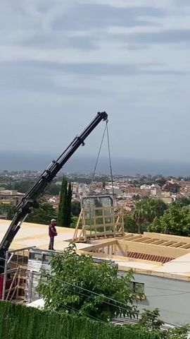A crane is lifting a large, framed glass panel onto the roof of a building under construction. A person in a red shirt and dark pants stands on the roof, seemingly supervising the operation. The building appears to be a multi-story structure with wooden framing visible. In the background, there is a city with terracotta roofs and trees, overlooking the ocean under a cloudy sky. Green foliage in the foreground partially obscures the view.