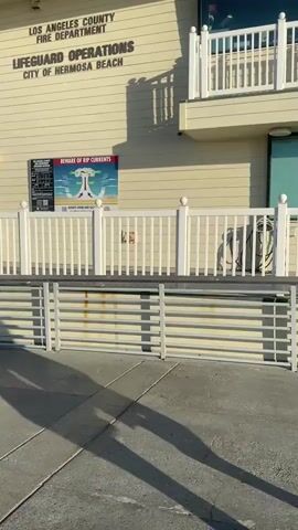 The video shows the exterior of a building labeled "LOS ANGELES COUNTY FIRE DEPARTMENT LIFEGUARD OPERATIONS CITY OF HERMOSA BEACH". The building is cream-colored with a white railing and a small balcony area. A sign with information about rip currents is posted on the wall. A shadow of a person is visible in the foreground on the pavement. The sky is clear and blue. The video pans slightly to the right, showing more of the building and some palm trees in the distance.