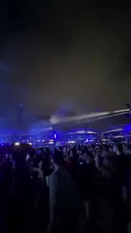 A large crowd of people is gathered at night for an outdoor event, likely a concert or festival. Above the crowd, bright blue lasers cut through the air, creating an atmosphere of excitement. A large screen displays a video of a man wearing sunglasses and a colorful hat, who seems to be performing or DJing, adding to the lively vibe. The people in the crowd are moving and appear to be enjoying the show. The night sky is visible overhead, enhancing the sense of being at a large-scale outdoor event.
