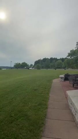 The video pans from a grassy field with trees in the distance, to a paved walkway alongside a stone wall. There is a informational kiosk next to the walkway, and further along, a picnic table with chairs. Beyond the kiosk, a parking lot is visible with cars and trucks. A building with an ornate roof can be seen in the background. The sky is overcast.