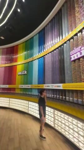 The video shows the inside of a store, possibly an M&M's store, with a large curved wall filled with transparent containers of M&M candies in a rainbow of colors. The containers are arranged with dispensers at the bottom for customers to fill bags. A yellow sign with "milk" is visible on the wall.  Below the dispensers is a decorative panel with white cut-out shapes.  The floor is made of light wood planks. People are seen walking around and looking at the candy. The price is listed as $1.69 per ounce.  Overhead are curved, linear lights.