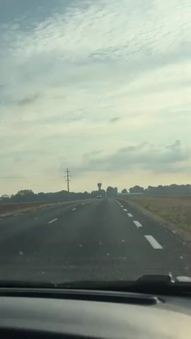 The video shows a first-person view from a car driving down a paved road. The road has white dashed lines on the right side and solid white lines on the left side indicating the lane. The road is straight with a slight incline ahead. The sky is overcast with scattered clouds. In the distance, there is a water tower and some trees. There are also power lines running along the side of the road. A white car is driving in the opposite direction on the road. The fields on the right side of the road appear to be harvested. The video is shot during the daytime.