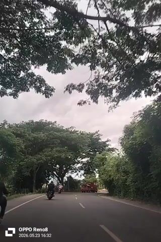 The video shows a road from the perspective of a vehicle driving on it. The road is lined with trees on both sides, creating a canopy effect. The sky is overcast with grey clouds. Two motorcycles are driving ahead on the same road, and a yellow truck, a red vehicle and a white car can be seen further down the road. The timestamp on the video indicates it was taken on May 23, 2026, at 16:32. The video is slightly shaky.